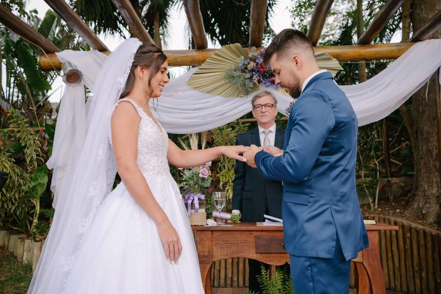 Fotografia de casamento em Jacareí: troca de alianças no altar da Chácara Lopes. O noivo Matthaus coloca a aliança na mão da noiva Rebeca sob o pergolado rústico, em um momento solene da cerimônia ao ar livre cercada pela natureza e luz do dia.