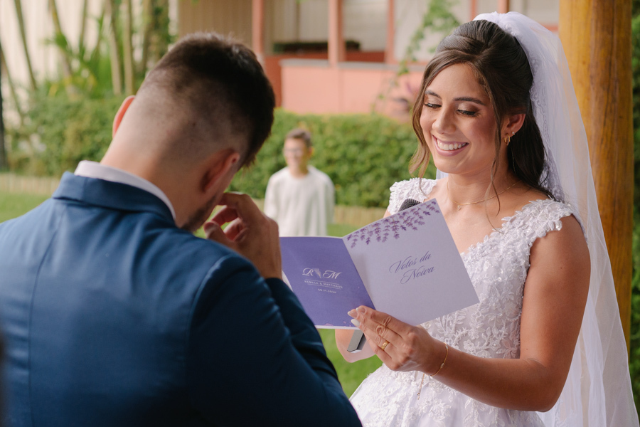 Fotografia de casamento em Jacareí: noiva Rebeca lê seus votos na Chácara Lopes. Ela sorri emocionada segurando o livreto de votos, enquanto o noivo Matthaus chora ao fundo. Registro sensível de cerimônia ao ar livre sob a luz natural do entardecer.