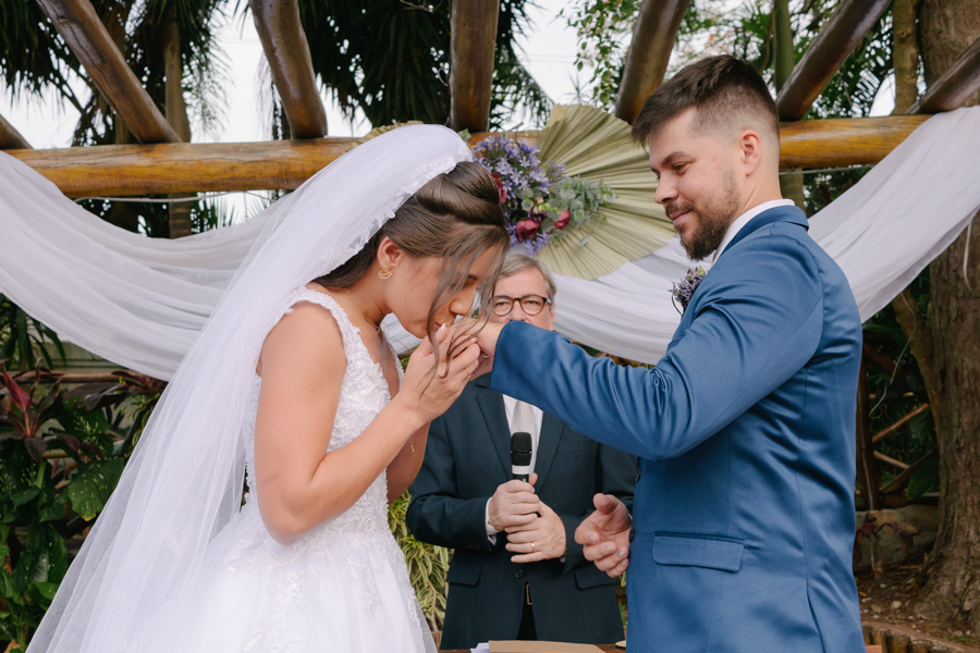 Fotografia de casamento em Jacareí: noiva Rebeca beija a mão do noivo Matthaus no altar da Chácara Lopes. Registro emocionante de carinho e respeito durante a cerimônia ao ar livre, sob o pergolado rústico e a luz natural do entardecer.