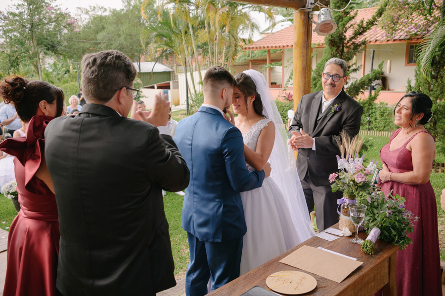 Fotografia de casamento em Jacareí: primeiro beijo de Matthaus e Rebeca no altar da Chácara Lopes. O casal se abraça sob o pergolado rústico enquanto os pais aplaudem emocionados, celebrando a união ao ar livre com a luz natural do fim de tarde.