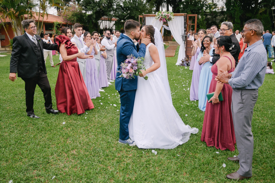 Fotografia de casamento em Jacareí: beijo dos noivos Matthaus e Rebeca após a cerimônia na Chácara Lopes. O casal celebra ao ar livre no gramado, cercado por padrinhos e convidados que aplaudem o momento sob a luz natural do entardecer.