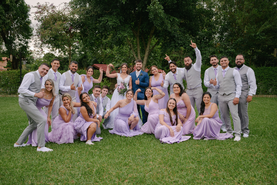 Fotografia de casamento em Jacareí: Matthaus e Rebeca com seus padrinhos na Chácara Lopes. Grande grupo posa animado no gramado ao ar livre, com madrinhas em tons de lavanda e padrinhos de colete cinza, celebrando a união com poses divertidas.