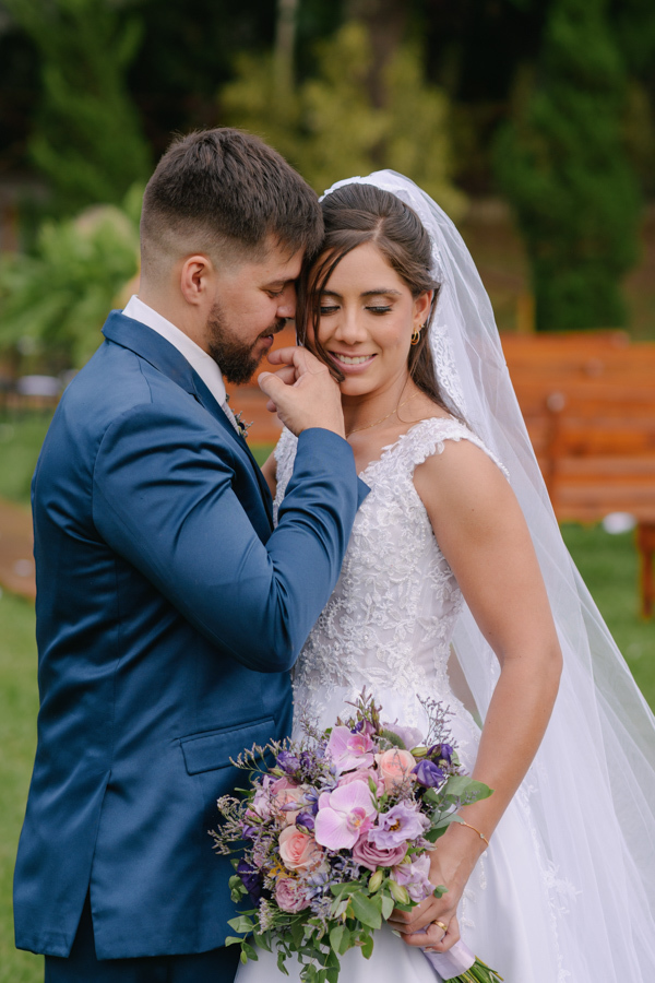 Fotografia de casamento em Jacareí: momento íntimo de Matthaus e Rebeca na Chácara Lopes. O noivo toca delicadamente o rosto da noiva, que sorri de olhos fechados. Registro romântico ao ar livre destacando o buquê de orquídeas e flores roxas.