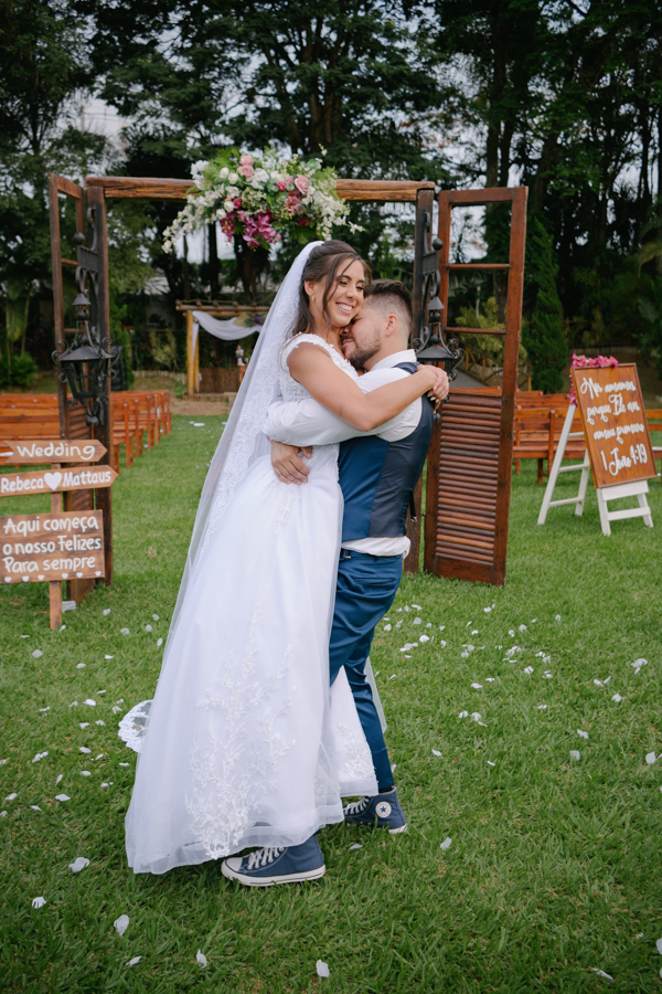 Fotografia de casamento em Jacareí: Matthaus levanta Rebeca em um abraço alegre na Chácara Lopes. O casal celebra no gramado em frente ao portal de madeira decorado com flores, com pétalas brancas espalhadas pelo chão sob a luz do entardecer.