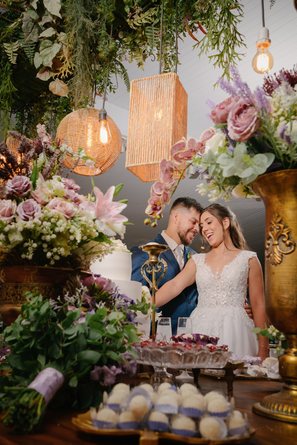 Fotografia de casamento em Jacareí: Matthaus e Rebeca na mesa do bolo na Chácara Lopes. Os noivos sorriem cercados por arranjos de flores roxas, doces finos e luminárias pendentes de fibra natural, em uma decoração rústica e sofisticada para a recepção.