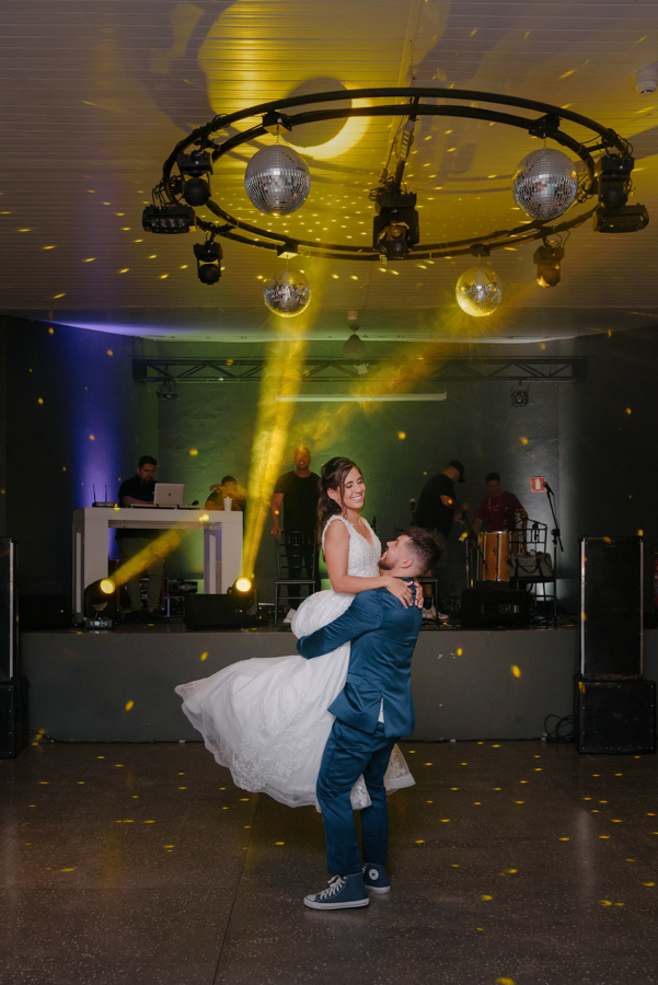 Fotografia de casamento em Jacareí: Matthaus ergue Rebeca em um abraço na pista de dança da Chácara Lopes. O casal celebra sob globos espelhados e luzes de festa, com o DJ e a banda ao fundo, em um momento vibrante e alegre da recepção.