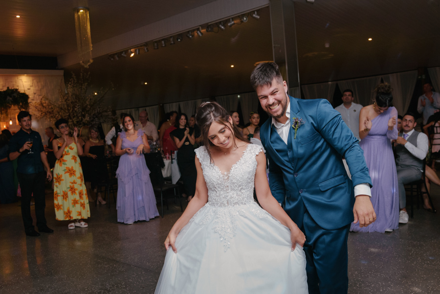 Fotografia de casamento em Jacareí: Matthaus e Rebeca se divertem na pista de dança da Chácara Lopes. Os noivos sorriem e fazem poses descontraídas, com a noiva segurando a saia do vestido e o noivo inclinado, celebrando com alegria junto aos convidados.