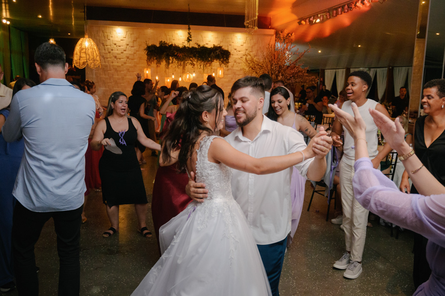 Fotografia de casamento em Jacareí: Matthaus e Rebeca dançam abraçados na pista de dança da Chácara Lopes. O casal sorri enquanto convidados aplaudem ao redor, em um ambiente festivo com iluminação quente e decoração rústica ao fundo.