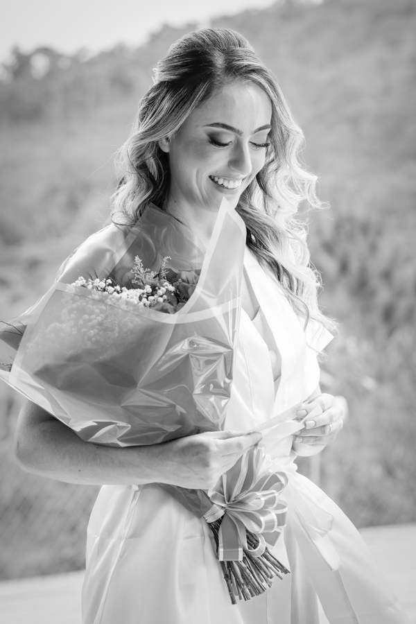 Noiva Aline sorrindo emocionada ao ler carta e segurar buquê de flores durante o making of na Chácara Mariah. Fotografia de casamento em Jacareí em preto e branco, destacando a sensibilidade e os momentos reais registrados por fotógrafo profissional.