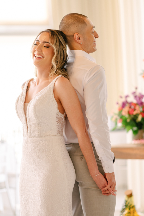 Noiva Aline e noivo Luidgi sorrindo de costas um para o outro e de mãos dadas na Chácara Mariah. Fotografia de casamento em Jacareí capturando a alegria e conexão do casal. Registro profissional por fotógrafo de casamento em Jacareí.