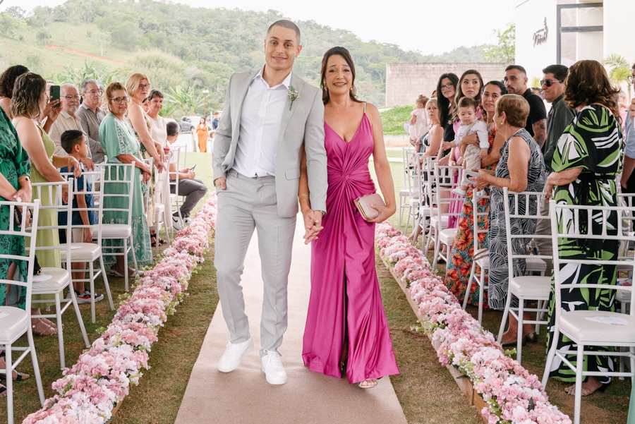 Entrada do noivo Luidgi acompanhado por sua mãe na cerimônia ao ar livre na Chácara Mariah. Fotografia de casamento em Jacareí registrando o caminho ao altar com convidados e decoração floral. Trabalho profissional de fotógrafo de casamento em Jacareí.