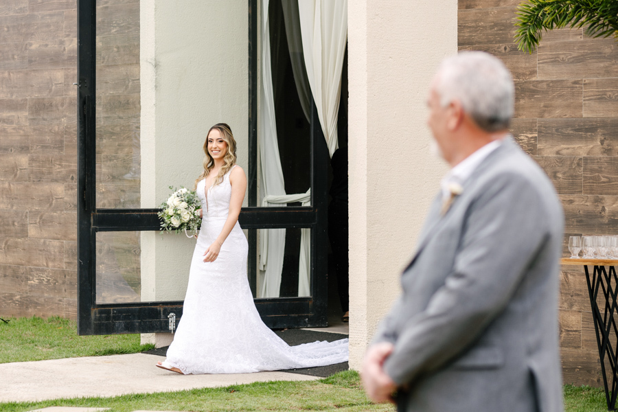 Entrada da noiva Aline na cerimônia ao ar livre na Chácara Mariah, em Jacareí. Fotografia de casamento capturando o sorriso da noiva com buquê e o pai em primeiro plano. Registro profissional por fotógrafo de casamento em Jacareí.
