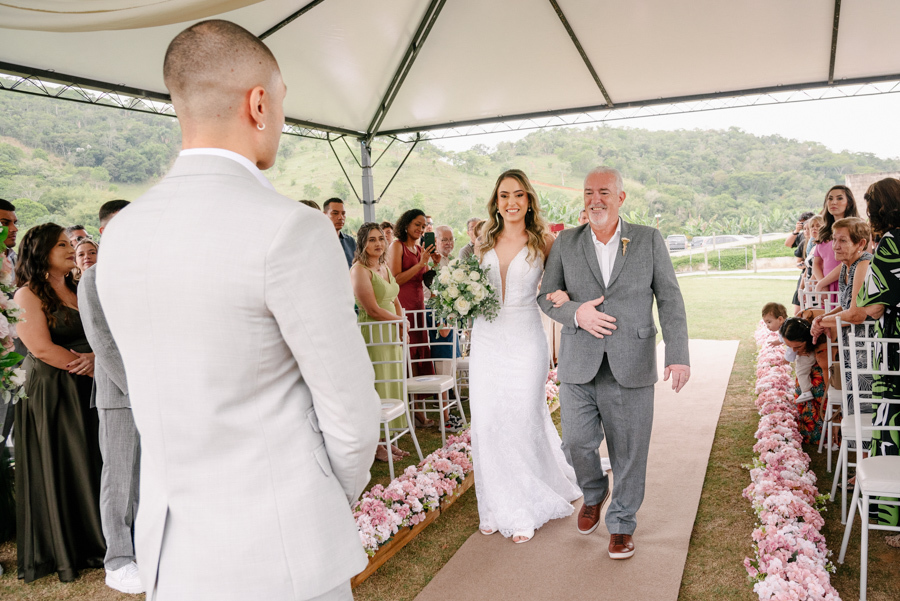 Noiva Aline entrando na cerimônia na Chácara Mariah, em Jacareí, acompanhada pelo pai sob o olhar do noivo Luidgi. Fotografia de casamento ao ar livre capturando a emoção do encontro no altar. Registro profissional por fotógrafo de casamento em Jacareí.