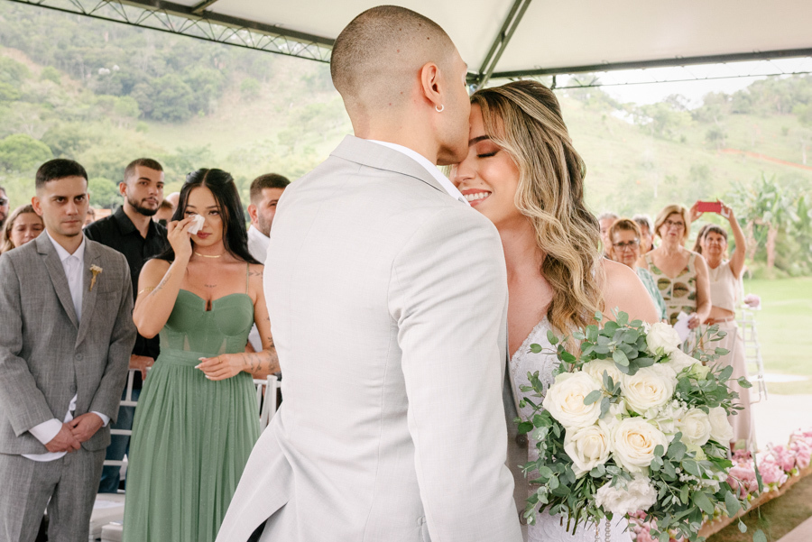 Noivo Luidgi beijando a testa da noiva Aline no altar na Chácara Mariah, em Jacareí. Fotografia de casamento capturando momento de carinho e emoção com convidados ao fundo. Registro profissional por fotógrafo de casamento em Jacareí.