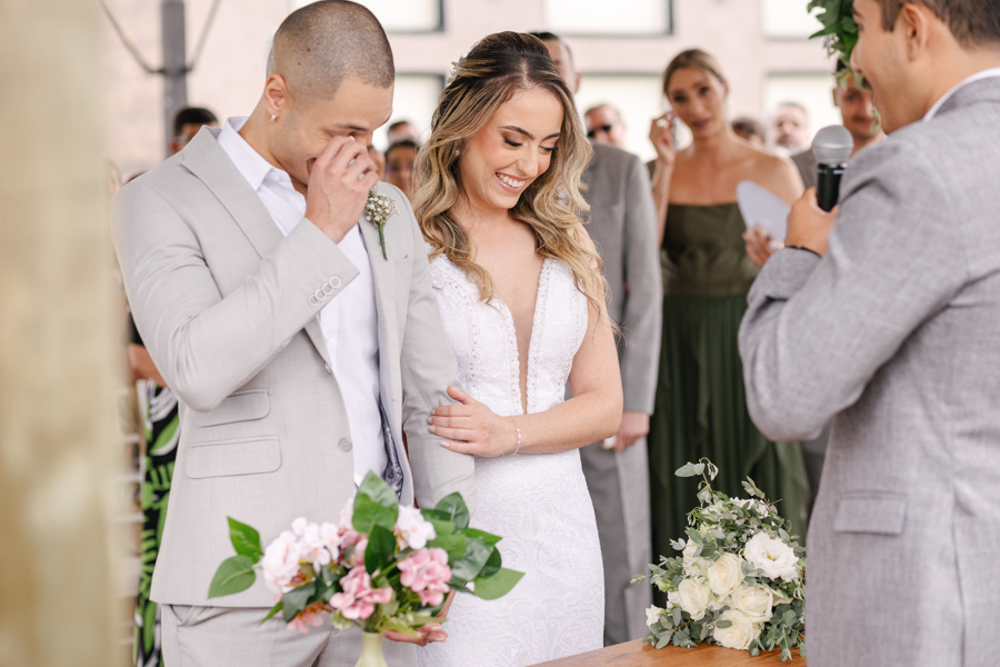 Noivo Luidgi emocionado secando lágrimas durante os votos no altar da Chácara Mariah, em Jacareí. Noiva Aline sorri ao seu lado em momento de profunda conexão. Fotografia de casamento em Jacareí registrando sentimentos reais por fotógrafo profissional.