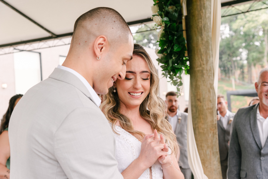 Noivos Aline e Luidgi sorrindo emocionados de mãos dadas no altar durante cerimônia na Chácara Mariah, em Jacareí. Fotografia de casamento capturando a felicidade e conexão do casal. Registro profissional por fotógrafo de casamento em Jacareí.