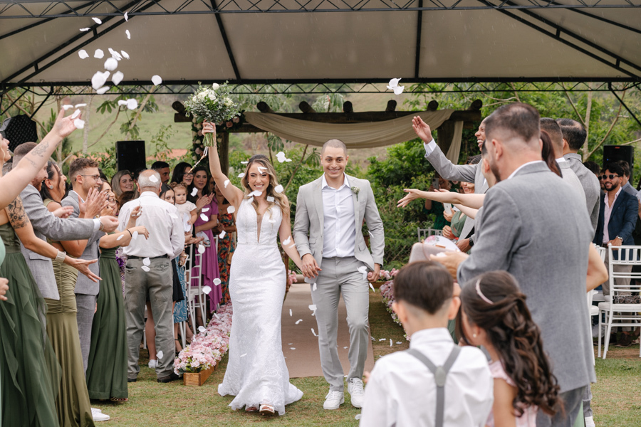 Saída dos noivos Aline e Luidgi sob chuva de pétalas brancas na Chácara Mariah, em Jacareí. Noiva celebra com buquê para o alto em momento de alegria após cerimônia ao ar livre. Fotografia profissional de casamento em Jacareí com momentos espontâneos.