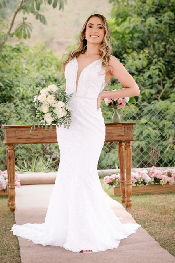 Retrato de corpo inteiro da noiva Aline sorrindo com seu buquê na Chácara Mariah, em Jacareí. Fotografia de casamento destacando o vestido sereia rendado e a decoração ao ar livre. Registro profissional realizado por fotógrafo de casamento em Jacareí.