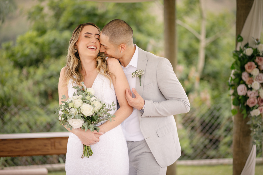 Noiva Aline sorrindo enquanto o noivo Luidgi a abraça por trás e beija seu ombro no altar da Chácara Mariah. Fotografia de casamento em Jacareí capturando um momento de leveza e afeto. Registro profissional realizado por fotógrafo de casamento em Jacareí.