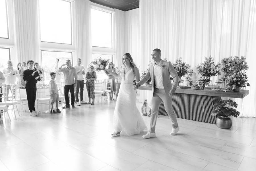 Entrada triunfal dos noivos Aline e Luidgi na recepção na Chácara Mariah, em Jacareí. Foto em preto e branco capturando a alegria do casal de mãos dadas, com convidados aplaudindo ao fundo. Registro profissional por fotógrafo de casamento em Jacareí.