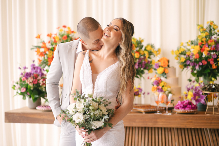 Noiva Aline sorrindo enquanto o noivo Luidgi beija seu pescoço em frente à mesa do bolo na Chácara Mariah. Fotografia de casamento em Jacareí destacando a alegria do casal e a decoração floral colorida. Registro profissional por fotógrafo em Jacareí.