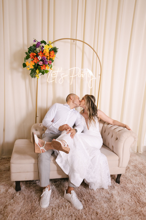 Noivos Aline e Luidgi se beijam sentados em um divã clássico sob letreiro de neon 