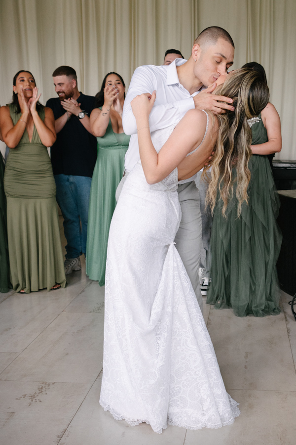 Noivo Luidgi inclinando e beijando a noiva Aline durante a festa na Chácara Mariah, em Jacareí. Fotografia de casamento capturando o beijo do casal sob aplausos das madrinhas ao fundo. Registro profissional realizado por fotógrafo de casamento em Jacareí.