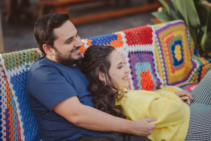 Abraço e cumplicidade: André e Nathália em ensaio pré-wedding em Campos do Jordão. Registro lifestyle aconchegante no sofá de crochê durante o outono na Serra da Mantiqueira. Fotógrafo de casamento capturando a essência e o carinho do casal.