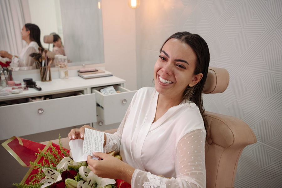 Noiva sorridente emocionada lendo carta e segurando buquê de flores vermelhas e brancas durante o making of. Fotografia de casamento em Jacareí na Chácara Mariah, destacando a emoção e os preparativos da Larissa para o grande dia.