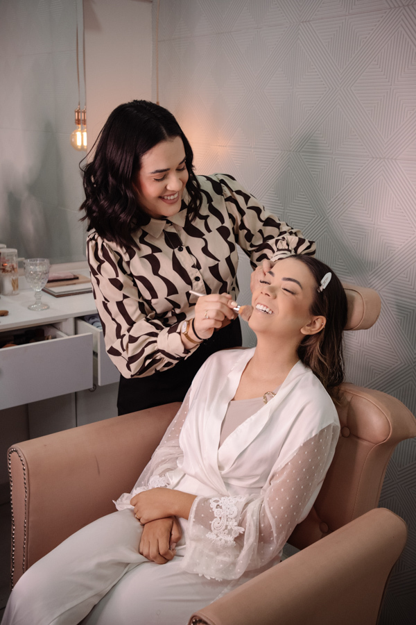 Making of da noiva Larissa em Jacareí: maquiadora aplica produtos enquanto a noiva sorri, sentada em poltrona rose. Fotografia de casamento profissional capturando a alegria e os preparativos na Chácara Mariah, com foco em momentos espontâneos e reais.