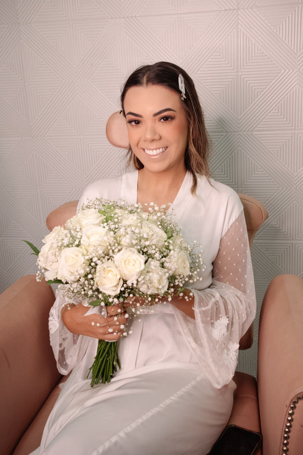 Noiva sorridente segurando buquê de rosas brancas e gipsofilas durante o making of em Jacareí. Fotografia de casamento profissional na Chácara Mariah, capturando a elegância e a expectativa da Larissa antes da cerimônia ao ar livre.