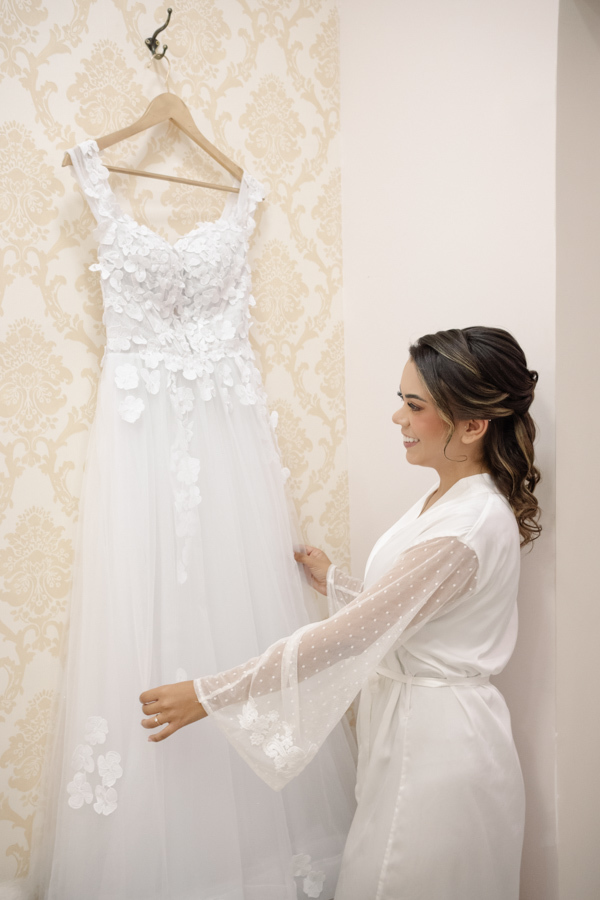 Noiva Larissa sorri admirando seu vestido de casamento branco com aplicações florais pendurado na parede. Fotografia profissional de casamento em Jacareí, capturando a emoção e os detalhes do making of na Chácara Mariah antes da cerimônia.