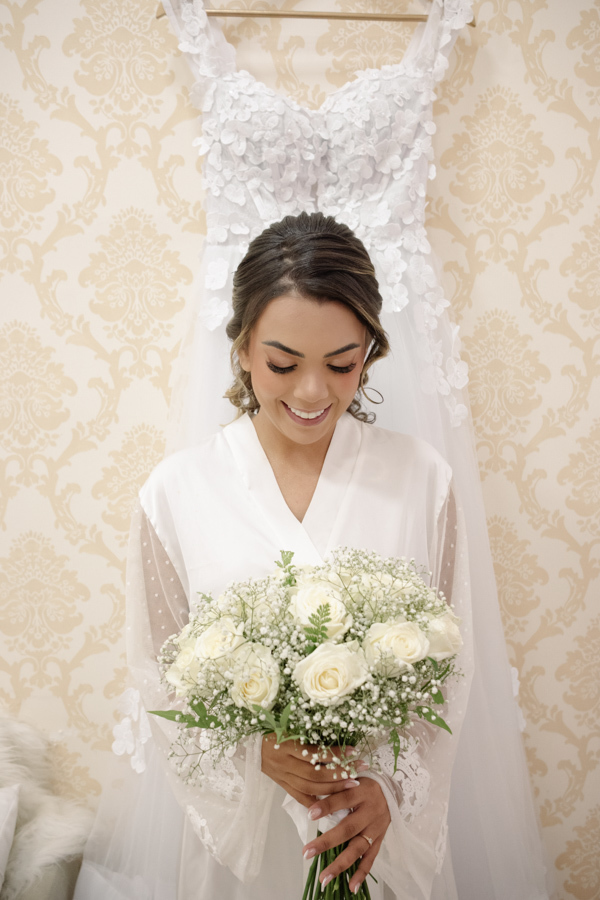 Noiva Larissa sorrindo, segurando buquê de rosas brancas à frente de seu vestido de noiva rendado. Fotografia profissional de casamento em Jacareí na Chácara Mariah, registrando a beleza e os detalhes finais do making of antes da cerimônia.