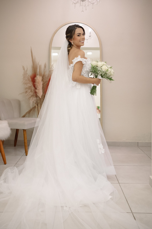 Noiva Larissa posa de costas exibindo o longo véu e o vestido de noiva com decote ombro a ombro e aplicações florais. Fotografia profissional de casamento em Jacareí na Chácara Mariah, capturando a elegância da noiva pronta para a cerimônia.
