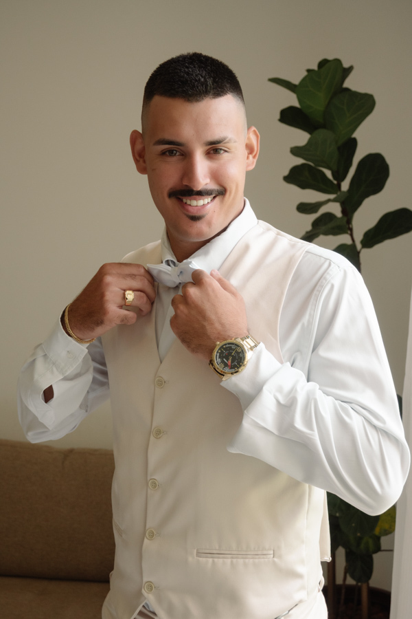 Noivo Ricardo sorridente durante o making of em Jacareí, vestindo colete bege e ajustando a gravata borboleta branca. Fotografia de casamento na Chácara Mariah, capturando a elegância e a preparação do noivo para o grande dia.