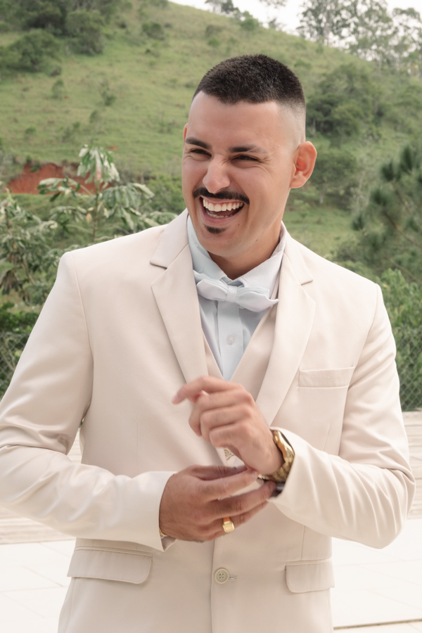 Noivo Ricardo sorrindo alegremente ao ar livre, vestindo terno bege e gravata borboleta. Fotografia profissional de casamento em Jacareí na Chácara Mariah, capturando a descontração e felicidade do noivo em meio à natureza antes da cerimônia.