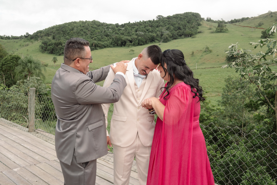 Noivo Ricardo emocionado recebe o carinho e a bênção dos pais ao ar livre na Chácara Mariah. Fotografia de casamento em Jacareí capturando um momento de profunda conexão familiar e afeto antes do início da cerimônia no campo.