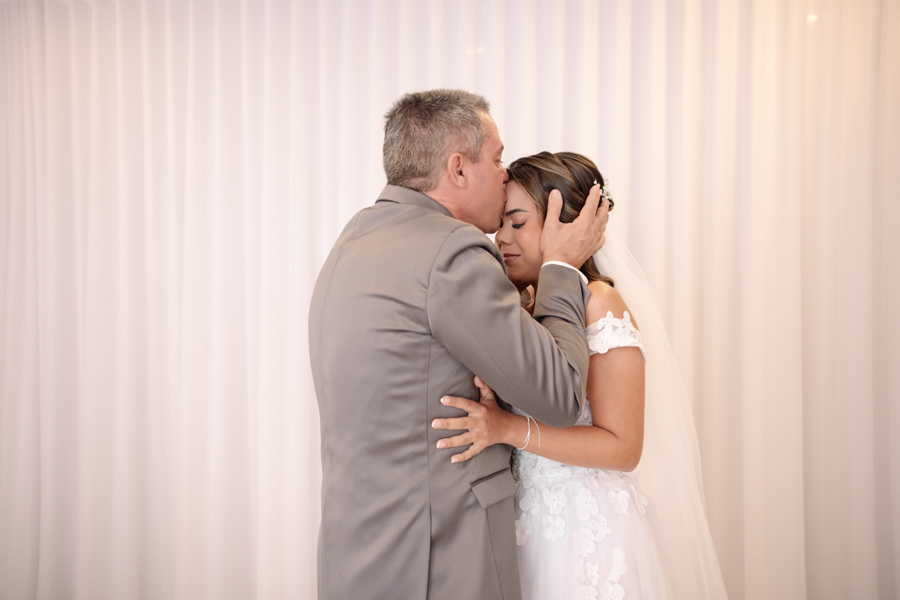 Pai beija a testa da noiva Larissa em um momento de profunda emoção durante o making of em Jacareí. Fotografia profissional de casamento na Chácara Mariah, capturando o carinho, a despedida e a conexão familiar antes da cerimônia ao ar livre.