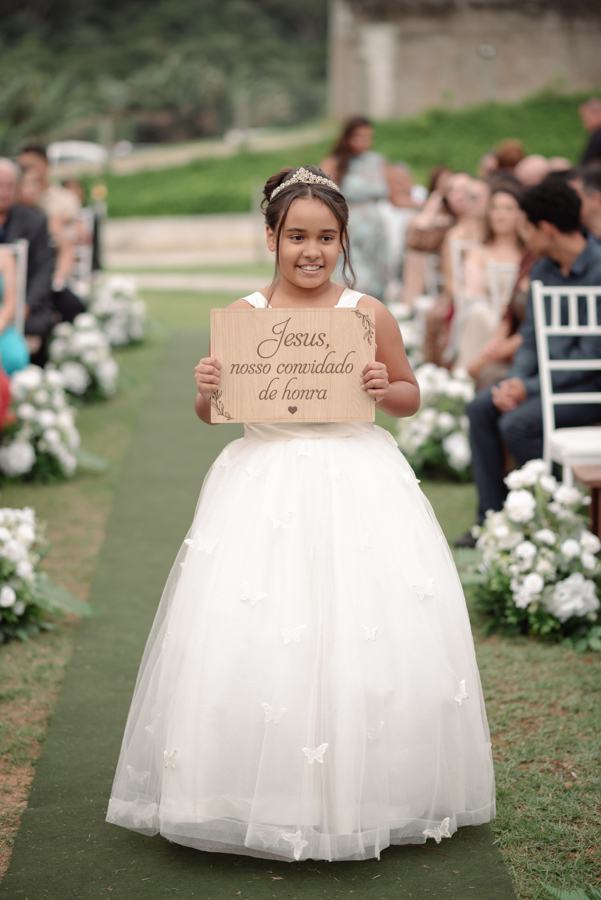 Daminha de honra entra na cerimônia de casamento ao ar livre na Chácara Mariah, em Jacareí. Ela usa vestido branco com borboletas e segura uma placa de madeira com a frase 