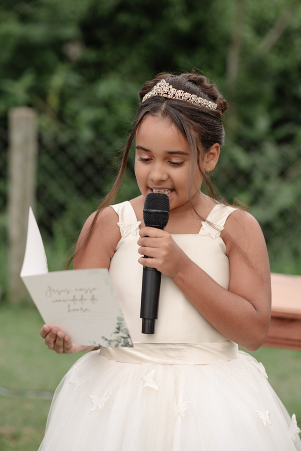 Daminha de honra em Jacareí lendo mensagem ao microfone durante cerimônia na Chácara Mariah. Fotografia de casamento profissional capturando a doçura e a participação das crianças em celebrações ao ar livre, com foco em detalhes e emoção espontânea.