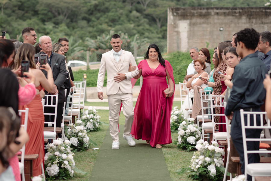 Entrada do noivo Ricardo com sua mãe em cerimônia ao ar livre na Chácara Mariah. Fotografia de casamento em Jacareí capturando o cortejo nupcial no gramado, com convidados acompanhando o momento sob a luz natural do Vale do Paraíba.