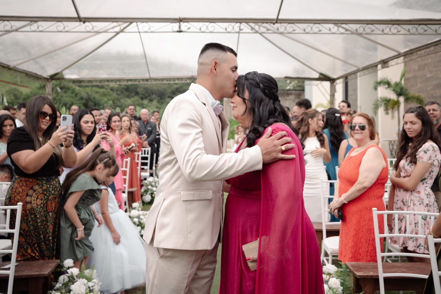 Noivo Ricardo beija a testa de sua mãe no altar em cerimônia ao ar livre na Chácara Mariah. Fotografia de casamento em Jacareí capturando a emoção, o respeito e o carinho familiar sob a tenda da celebração, com convidados ao fundo.