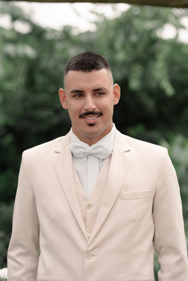 Noivo Ricardo posa sorridente ao ar livre vestindo terno bege e gravata borboleta branca. Fotografia de casamento em Jacareí na Chácara Mariah, destacando a elegância do noivo e a iluminação natural do Vale do Paraíba para o grande dia.