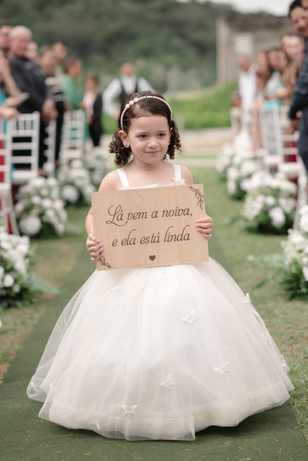 Daminha de honra em Jacareí caminhando pelo gramado da Chácara Mariah. Ela segura uma placa de madeira com a frase 