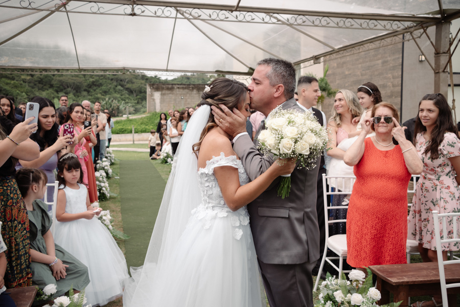 Pai beija a testa da noiva Larissa no altar sob a tenda da cerimônia ao ar livre na Chácara Mariah. Fotografia de casamento em Jacareí capturando a despedida emocionante, o carinho familiar e a entrega da noiva para o início da celebração.