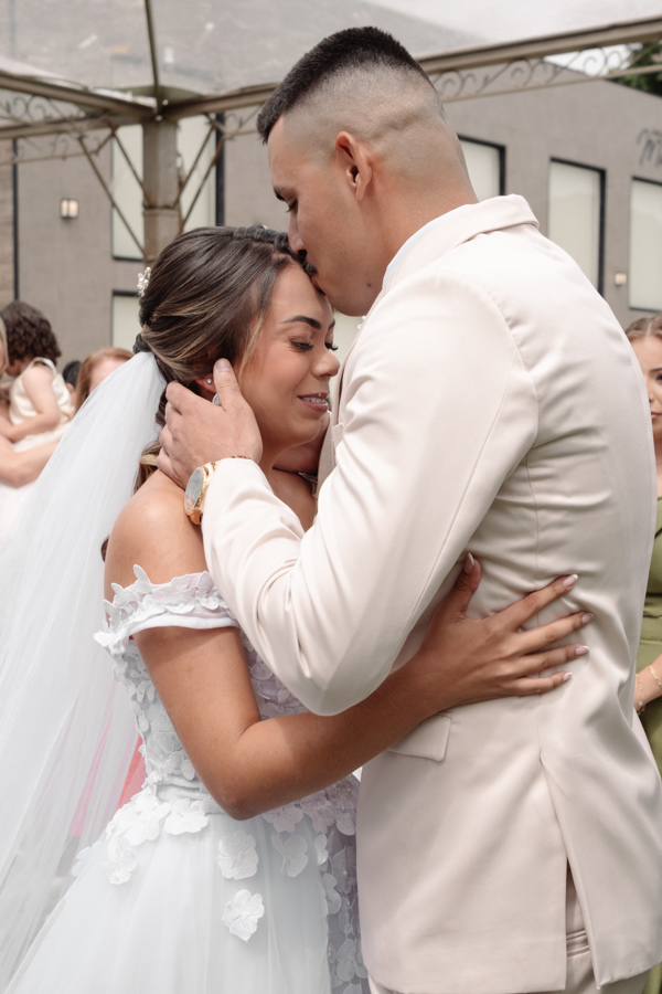 Noivo Ricardo beija a testa da noiva Larissa no altar, em um momento de profunda emoção durante a cerimônia ao ar livre. Fotografia profissional de casamento em Jacareí na Chácara Mariah, capturando o carinho e o início de uma nova vida juntos.