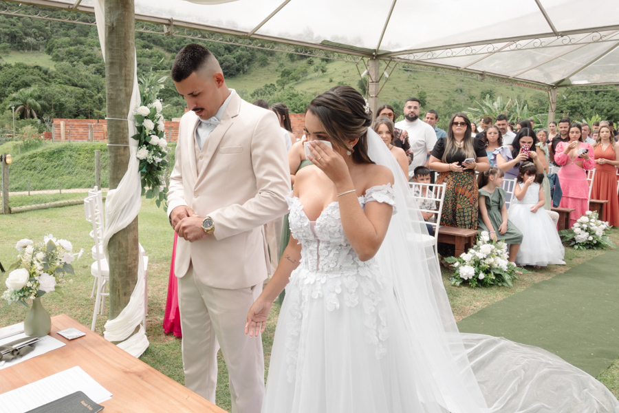 Noiva Larissa emocionada enxuga lágrimas no altar ao lado do noivo Ricardo durante cerimônia ao ar livre. Fotografia profissional de casamento em Jacareí na Chácara Mariah, capturando a intensidade e a sensibilidade do 