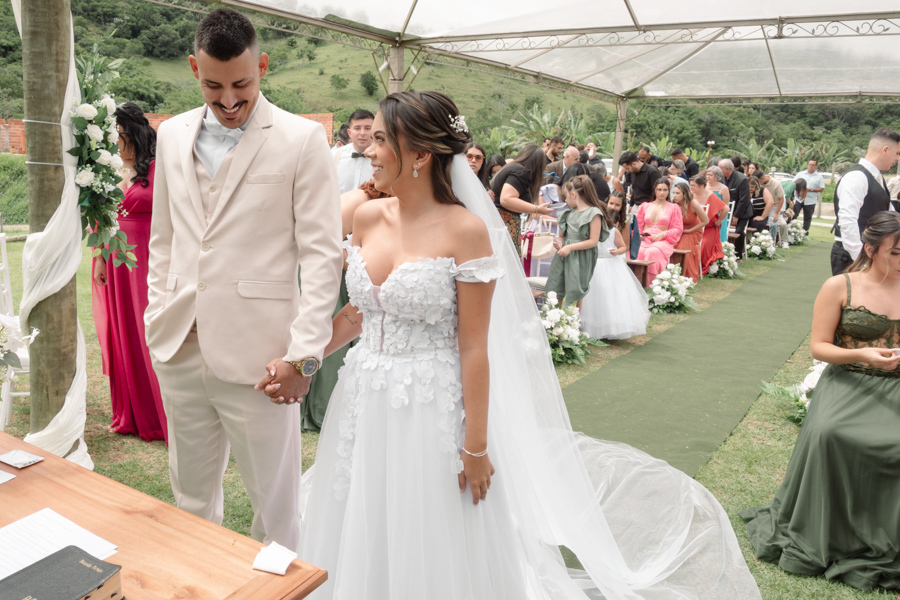 Noiva Larissa e noivo Ricardo de mãos dadas no altar durante cerimônia ao ar livre na Chácara Mariah, em Jacareí. Fotografia profissional capturando o casal sorridente sob a tenda, com convidados ao fundo e a paisagem verde do Vale do Paraíba.