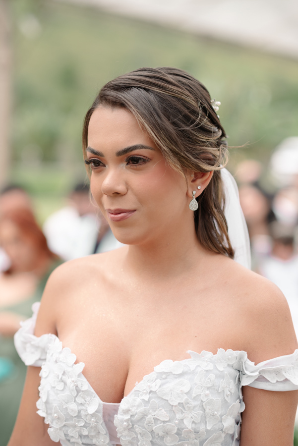 Close-up da noiva Larissa no altar durante cerimônia ao ar livre na Chácara Mariah, em Jacareí. Fotografia profissional destacando sua maquiagem clássica, penteado com acessórios de pérolas e os detalhes florais do vestido de noiva ombro a ombro.
