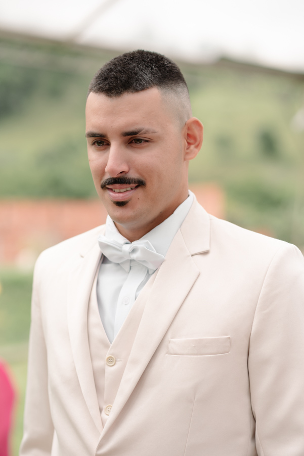 Noivo Ricardo no altar durante cerimônia ao ar livre na Chácara Mariah, em Jacareí. Fotografia profissional capturando o noivo sorridente, vestindo terno bege e gravata borboleta branca, com a paisagem verde do Vale do Paraíba ao fundo.